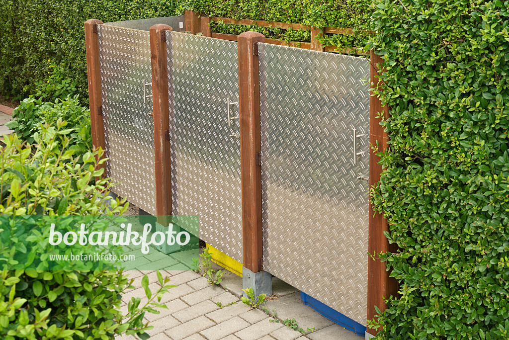 534256 - Modern litter bin with patterned stainless steel plates between wooden posts on a hedge