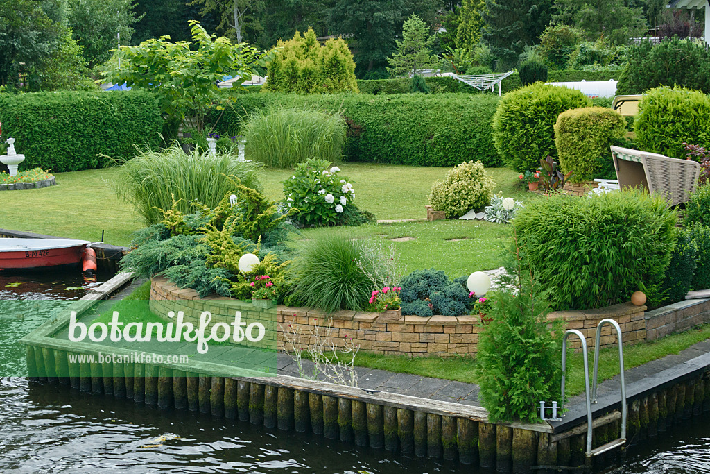 559033 - Well-kept garden with solitary plants, hedge, fountain, wall, wicker beach chair and boat on a water property