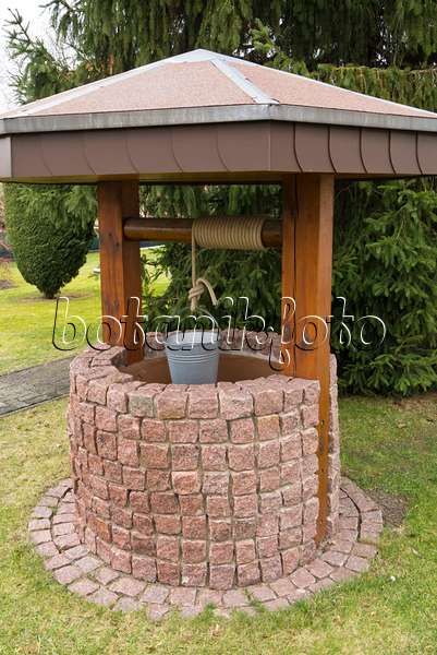 541004 - Aus Steinquadern gemauerter und überdachter Ziehbrunnen mit einem an einem Seil hängenden Eimer in einem Garten