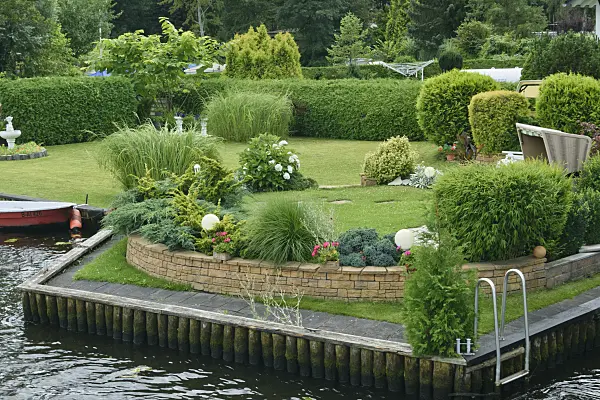 559033 - Gepflegter Garten mit Solitärpflanzen, Hecke, Brunnen, Mauer, Strandkorb und Boot auf einem Wassergrundstück
