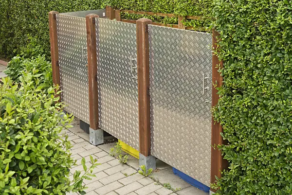 534256 - Poubelle moderne avec panneaux en acier inoxydable à motifs entre des poteaux en bois sur une haie
